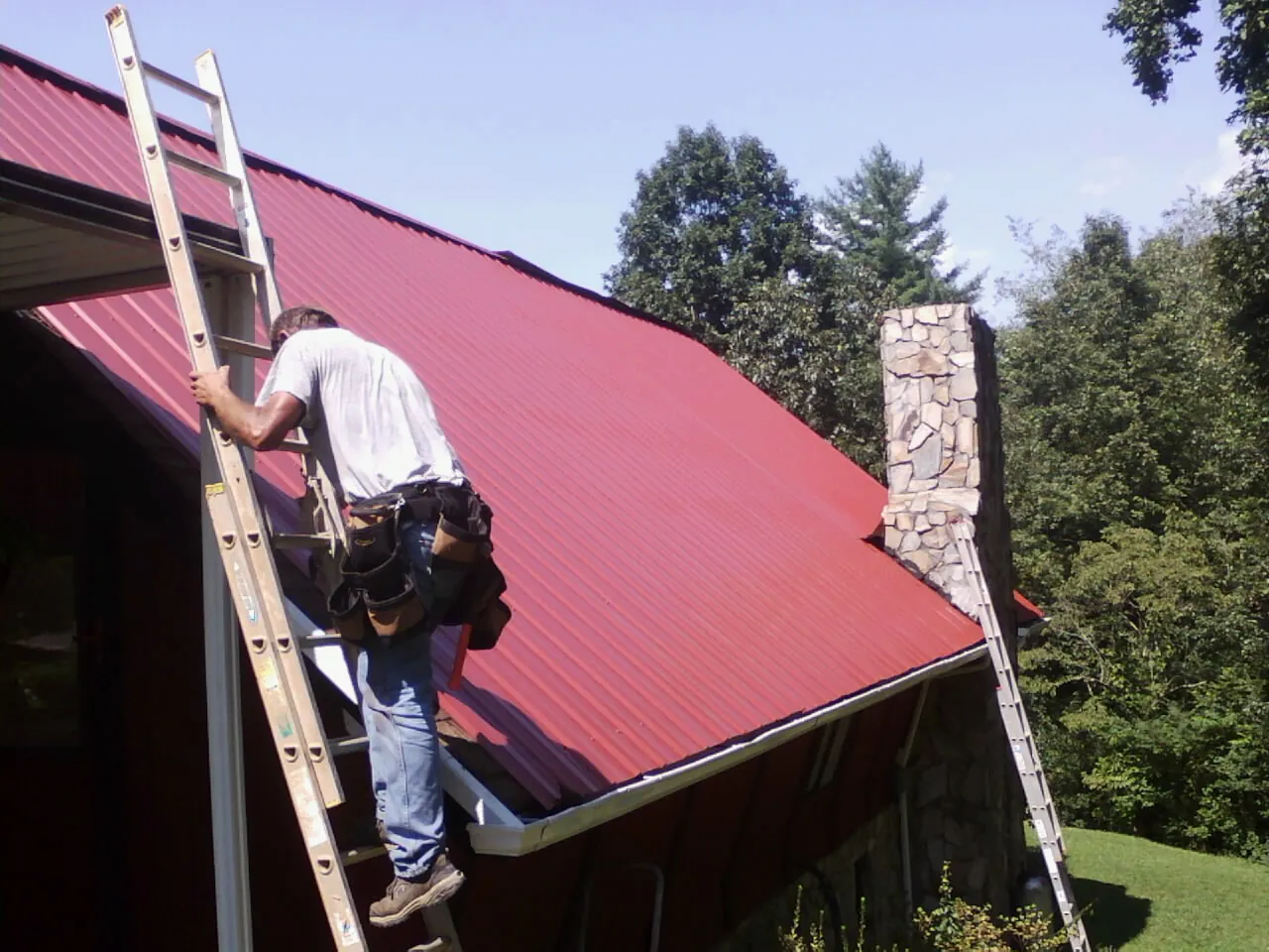 Roofer on a ladder beside a red metal roof and stone chimney — Soffit & Fascia Repair by Roofing Brothers in Cowiche